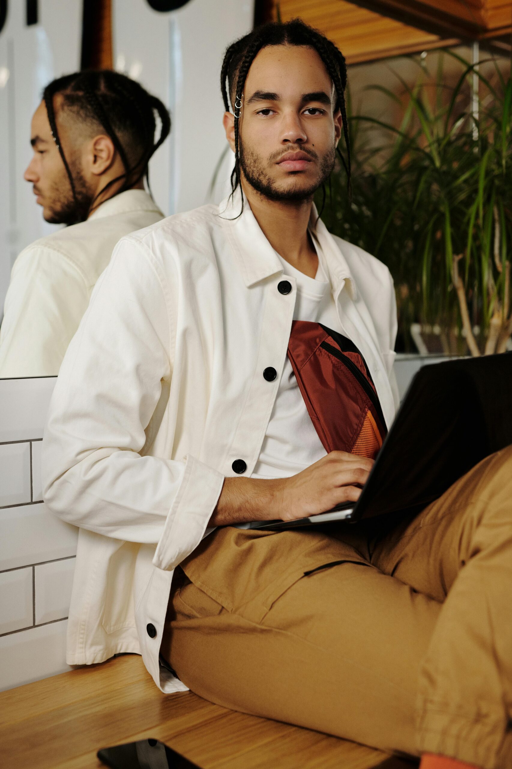 Stylish young adult man with braids using a laptop indoors with mirror reflection.