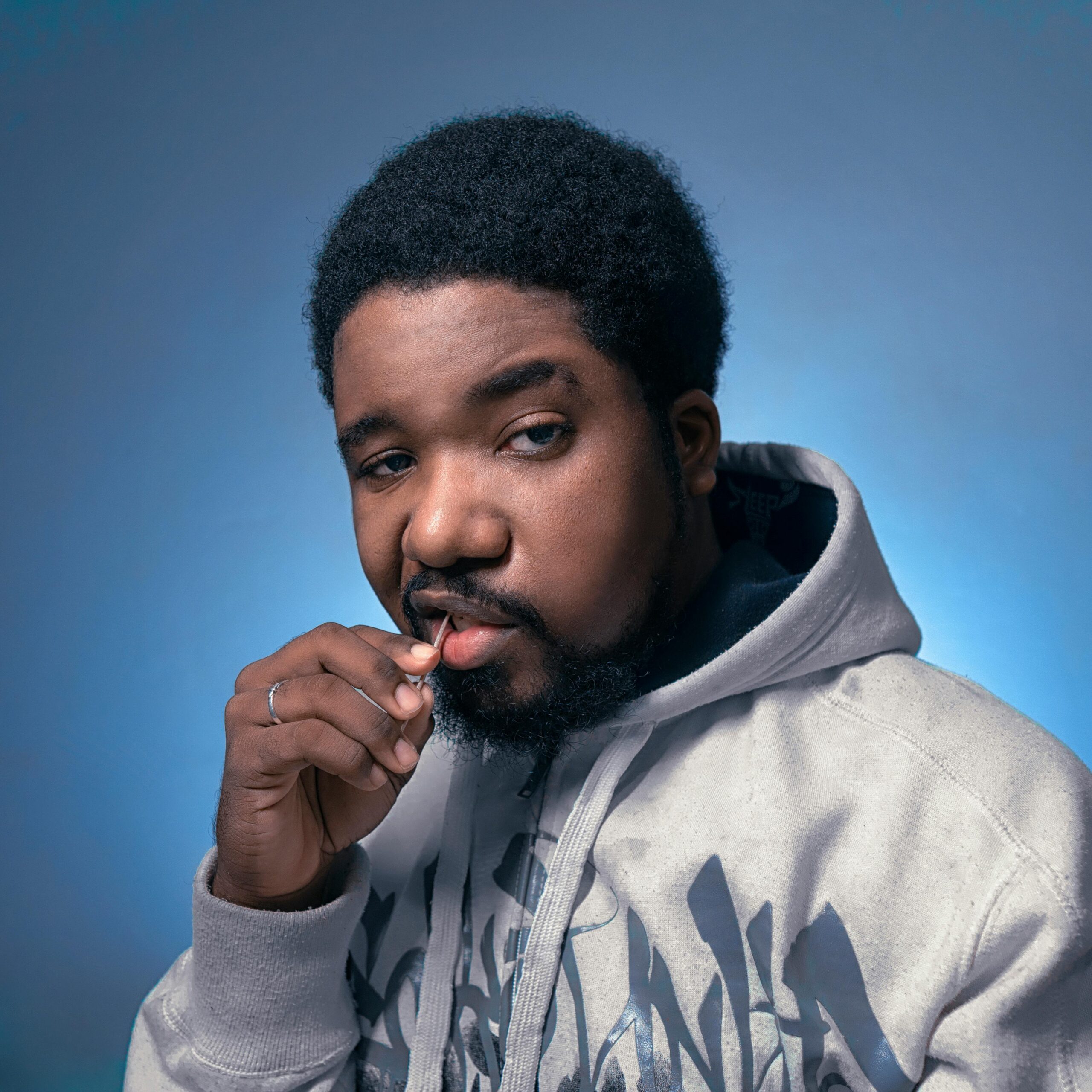 Stylish portrait of a man in a hoodie holding a toothpick against a blue background.